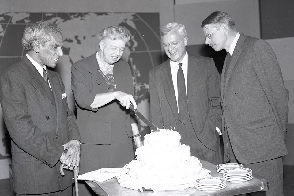 Eleanor Roosevelt stand with three other people and cuts a cake.