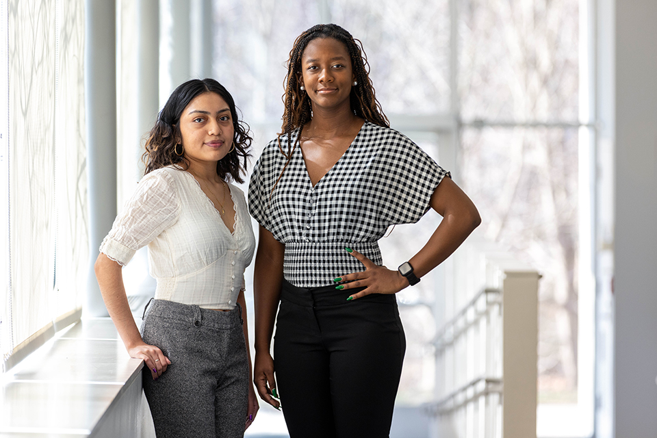 Two students with hands on hips stand in front of a window