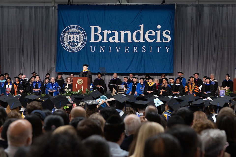 The backs of heads of commencement attendees are foreground for a giant blue and white Brandeis banner and the commencement dais