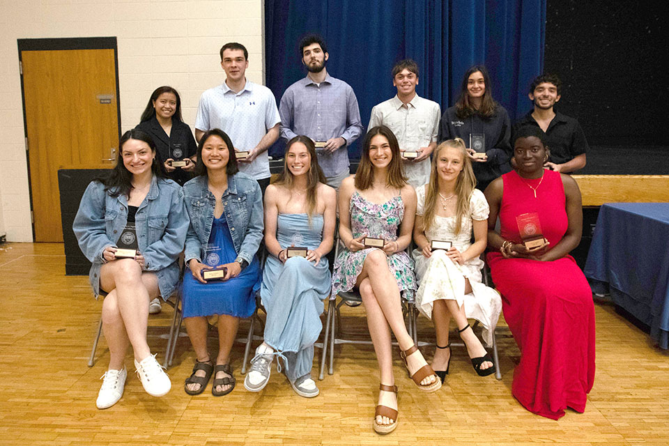 A group of student athletes sit in chairs and smile while holding their awards.