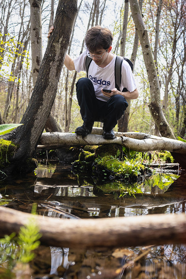 A student stands on a log over a stream in the woods.