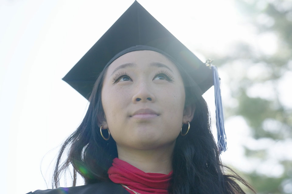 A student wearing a cap and gown looks up at the sky.