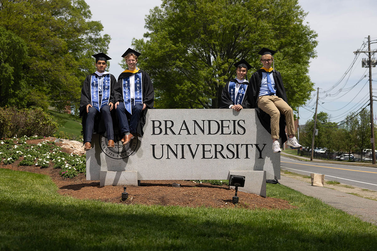 A group of students dressed in academic regalia sit on top of the Brandeis University sign.
