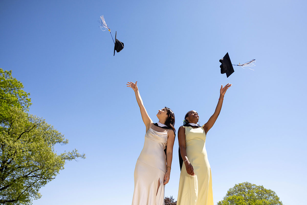 Two students stand on campus and throw their graduation caps in the air.