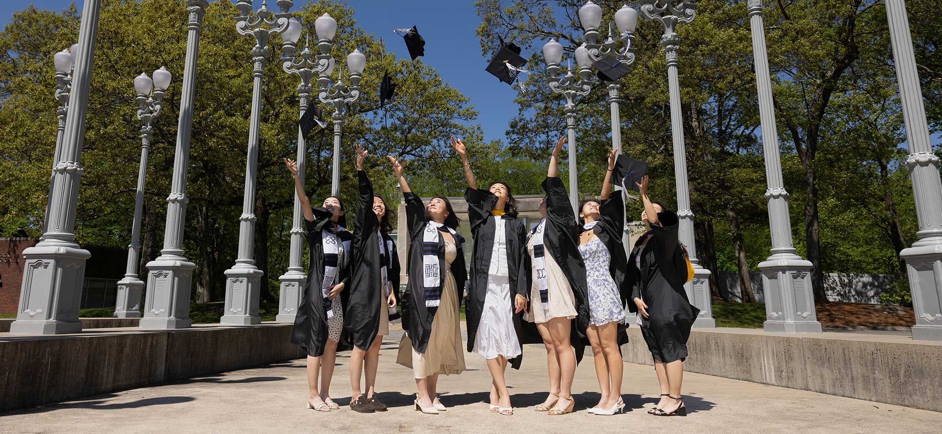 A group of graduates dressed in academic regalia throw their caps in the air at the "Light of Reason" sculpture.