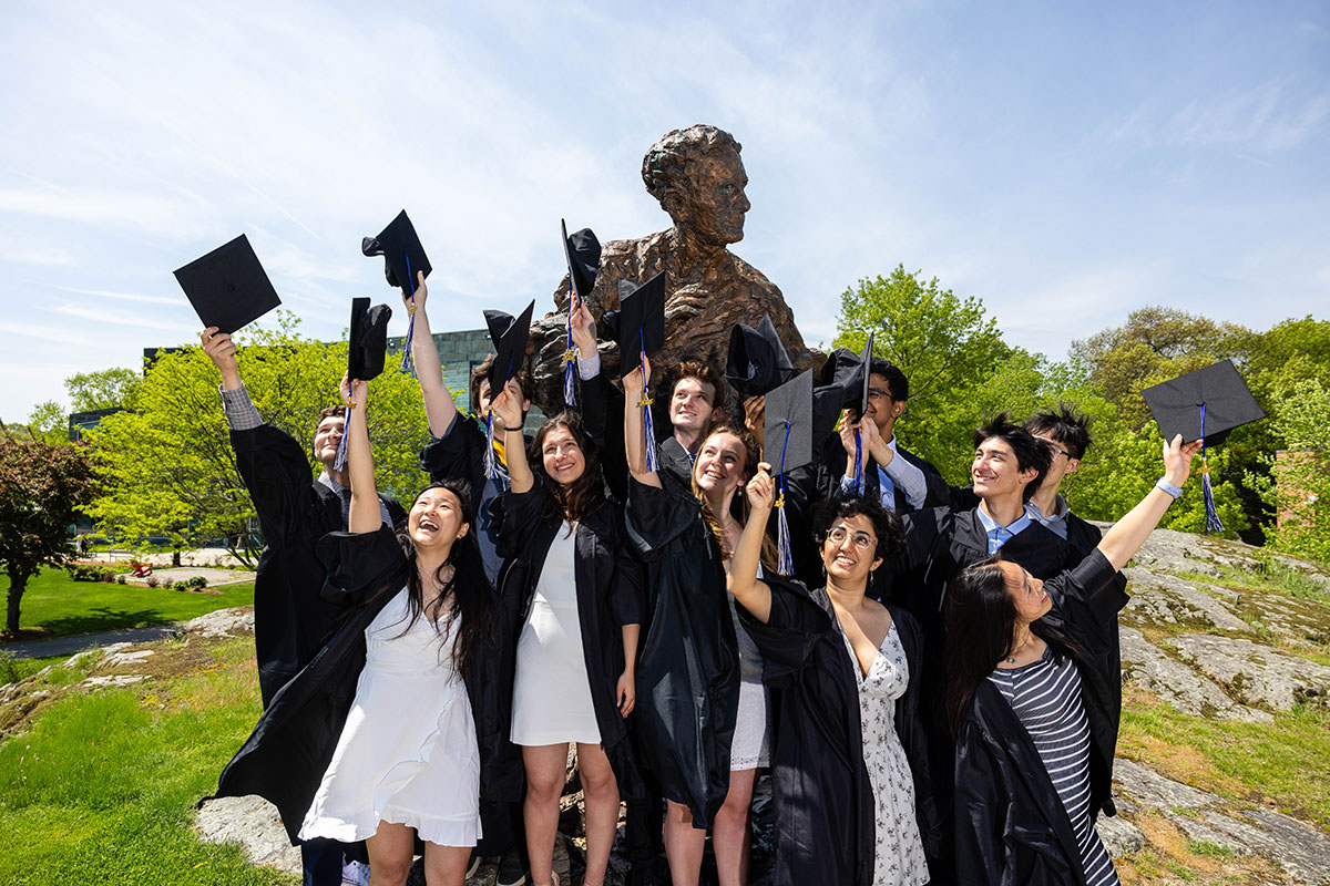 A group of graduates dressed in academic regalia stand in front of the Louis Brandeis statue and throw their caps in the air.