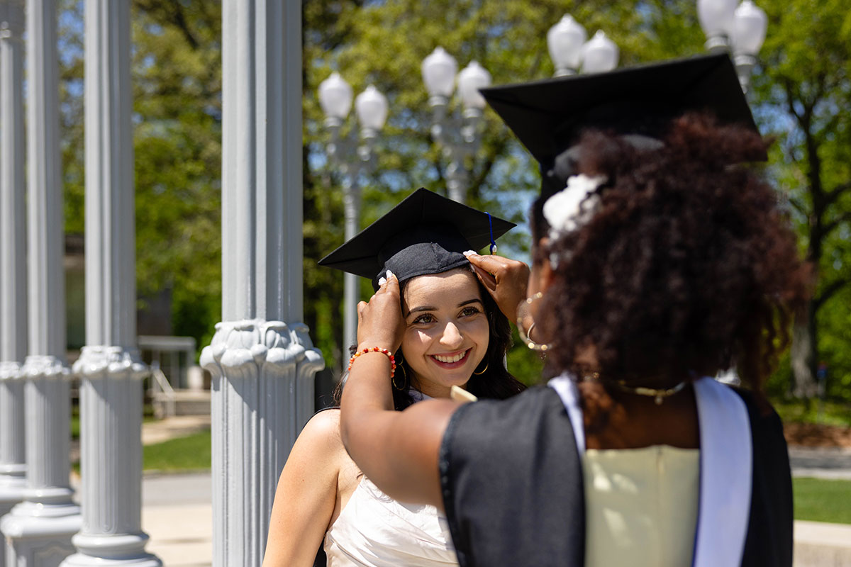 A student dressed in academic regalia adjusts her friend's cap.