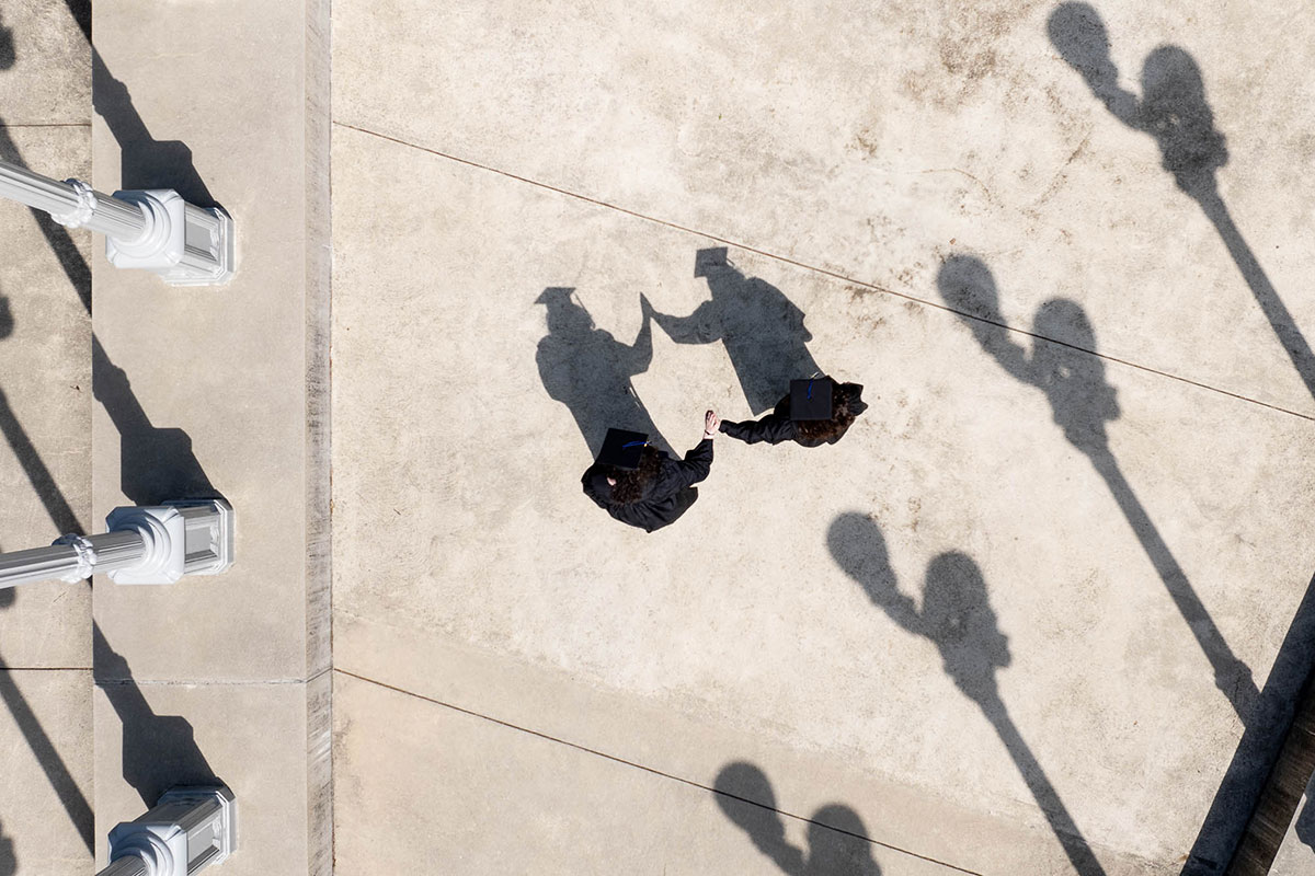 Aerial view of two students dressed in academic regalia high-fiving at the "Light of Reason" sculpture.