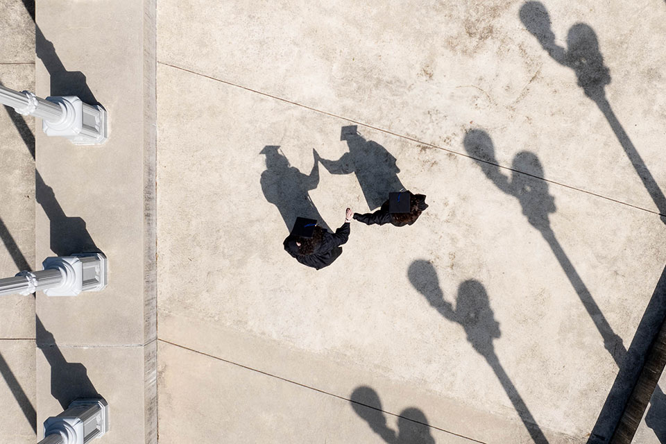 Aerial view of two students dressed in academic regalia high-fiving at the "Light of Reason" sculpture.