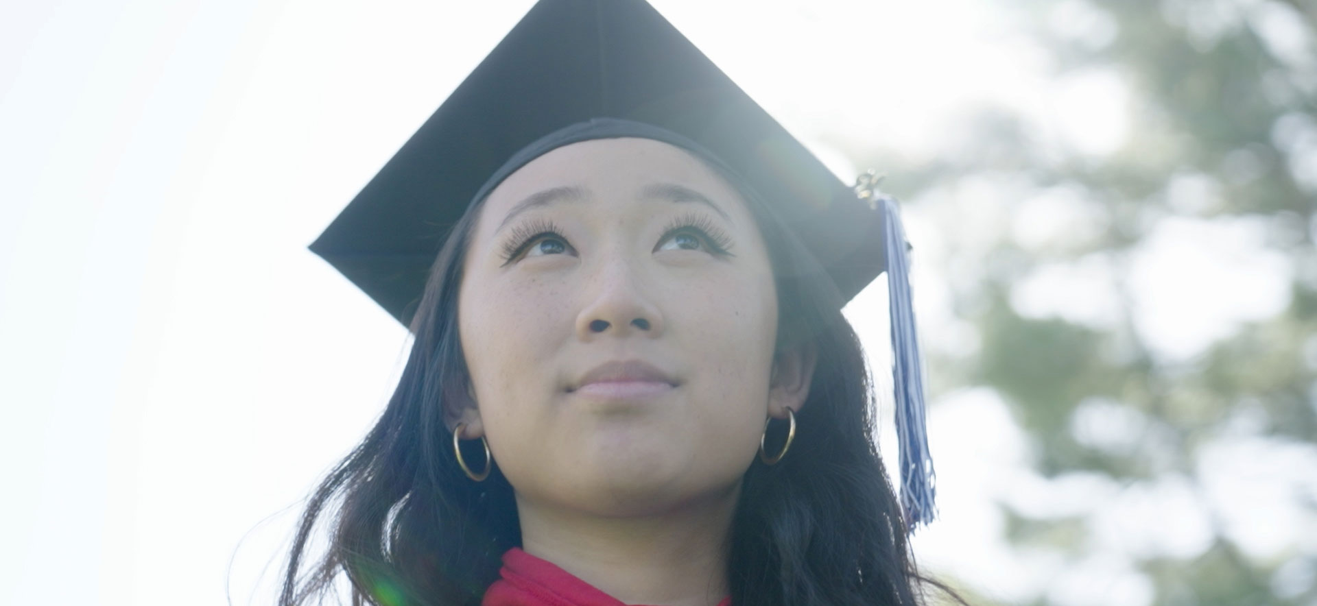 A student wearing a cap and gown looks up at the sky.
