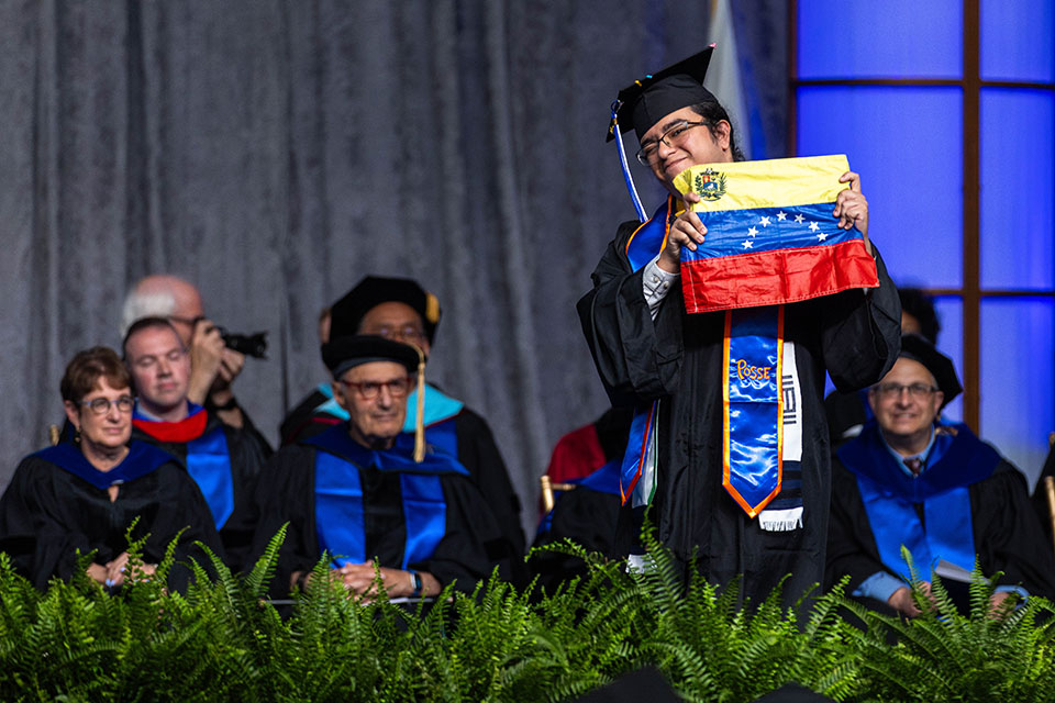 A student dressed in academic regalia smiles and holds up the Venezuelan flag on stage.