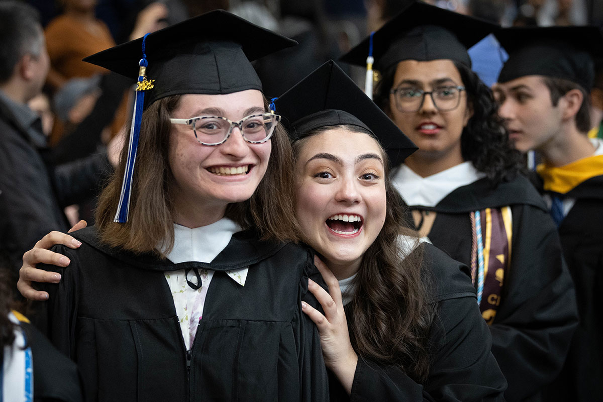 Two graduating students dressed in academic regalia smile at Commencement.