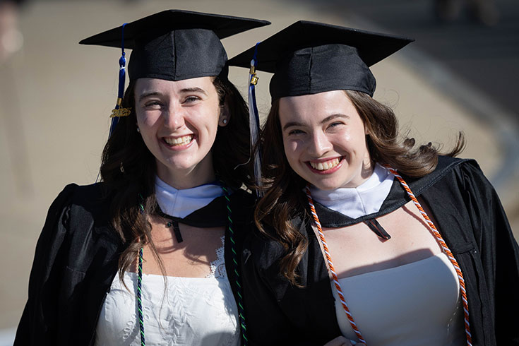 Two graduating students dressed in academic regalia smile outside on campus.