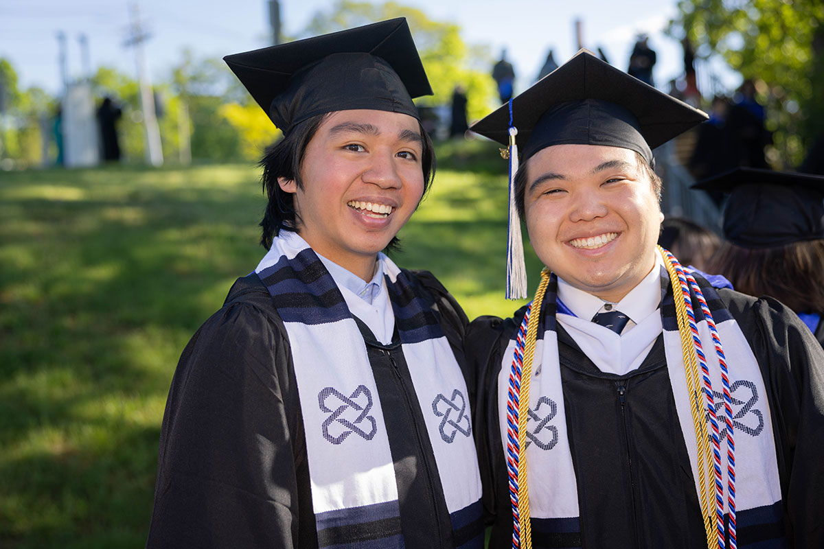 Two graduating students dressed in academic regalia smile together outside.