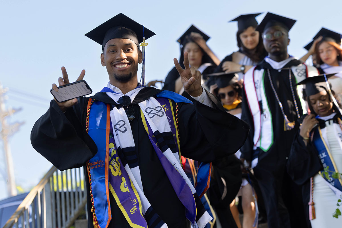 Graduating students walk down a flight of stairs wearing academic regalia. One holds up the peace sign.