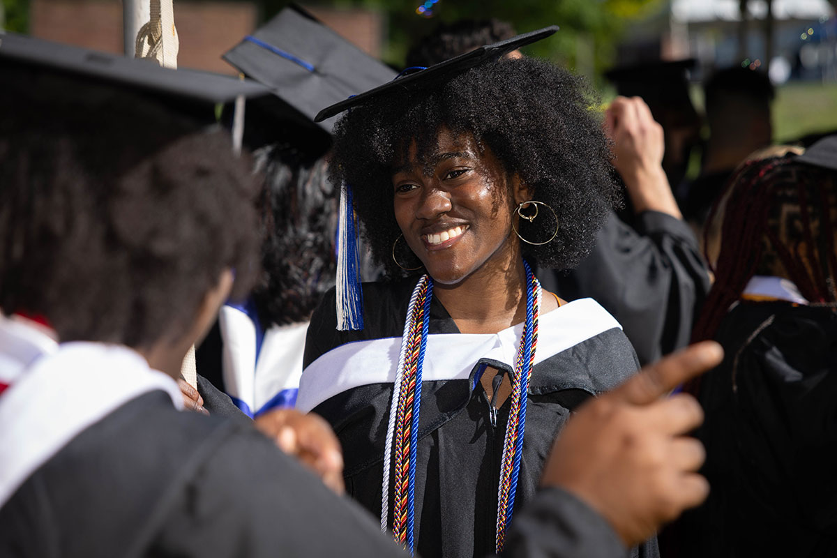 A graduating student dressed in academic regalia smiles at a friend.