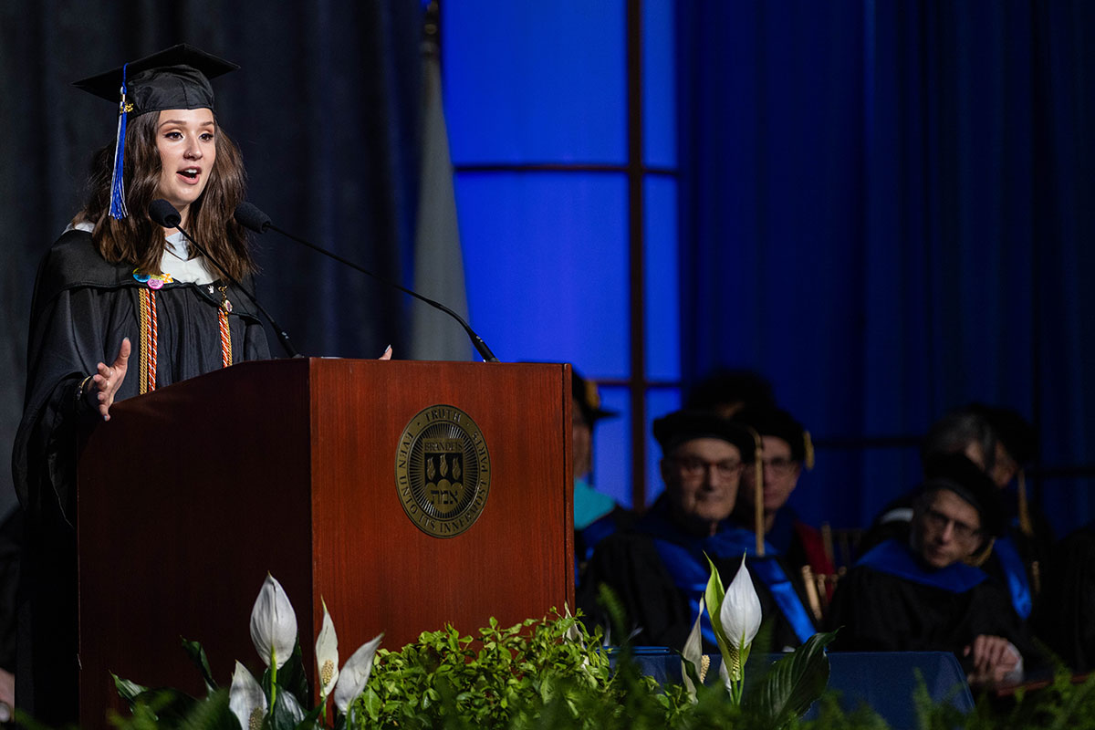 Hana Klempnauer Miller, dressed in academic regalia, addresses an audience from a podium at Commencement.