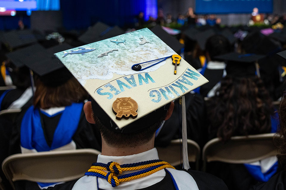 A graduating student wears a hand-decorated mortarboard with an image of a beach that reads "Making Waves."