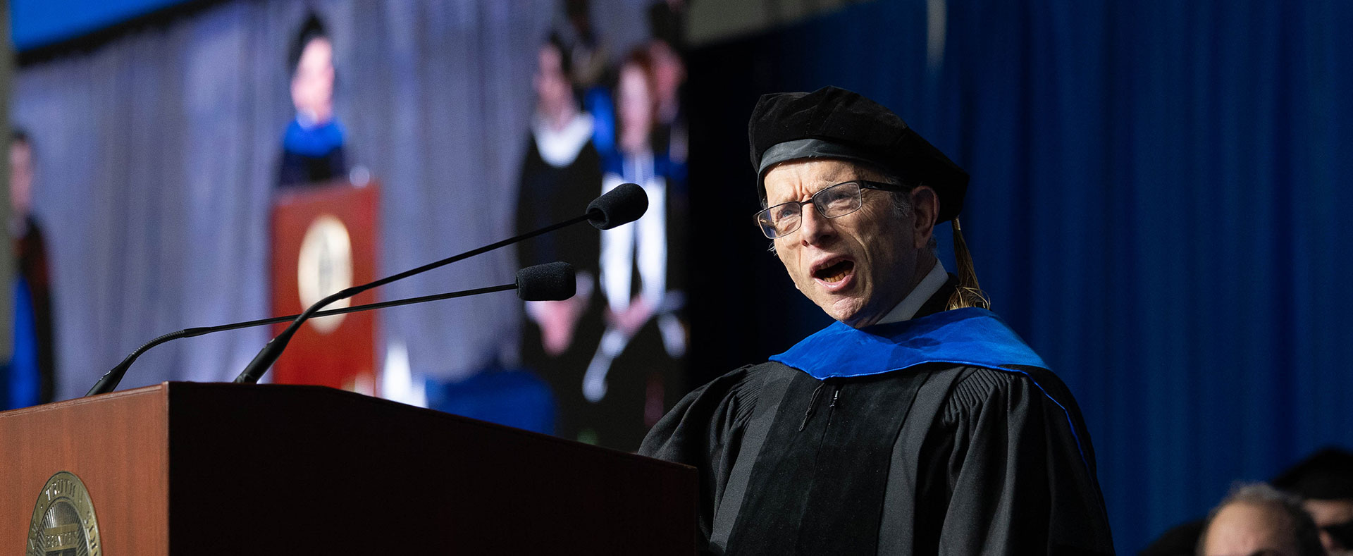 Jonathan Sarna, dressed in academic regalia, speaks into a microphone at a podium on stage at Commencement.