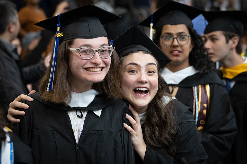 Two graduating students dressed in academic regalia smile at Commencement.
