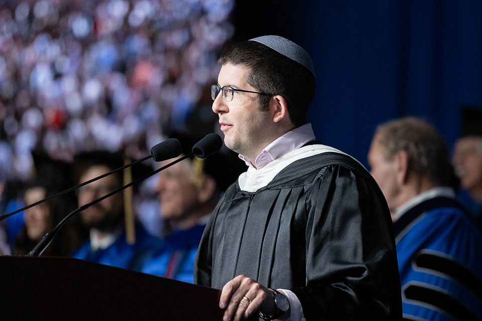Rabbi Seth Winberg speaks into a microphone at a podium on stage during Commencement.