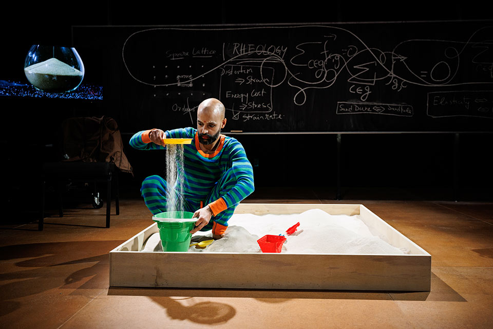 Shayok Misha Chowdhury crouches in a sandbox, watching sand sift into a green bucket.