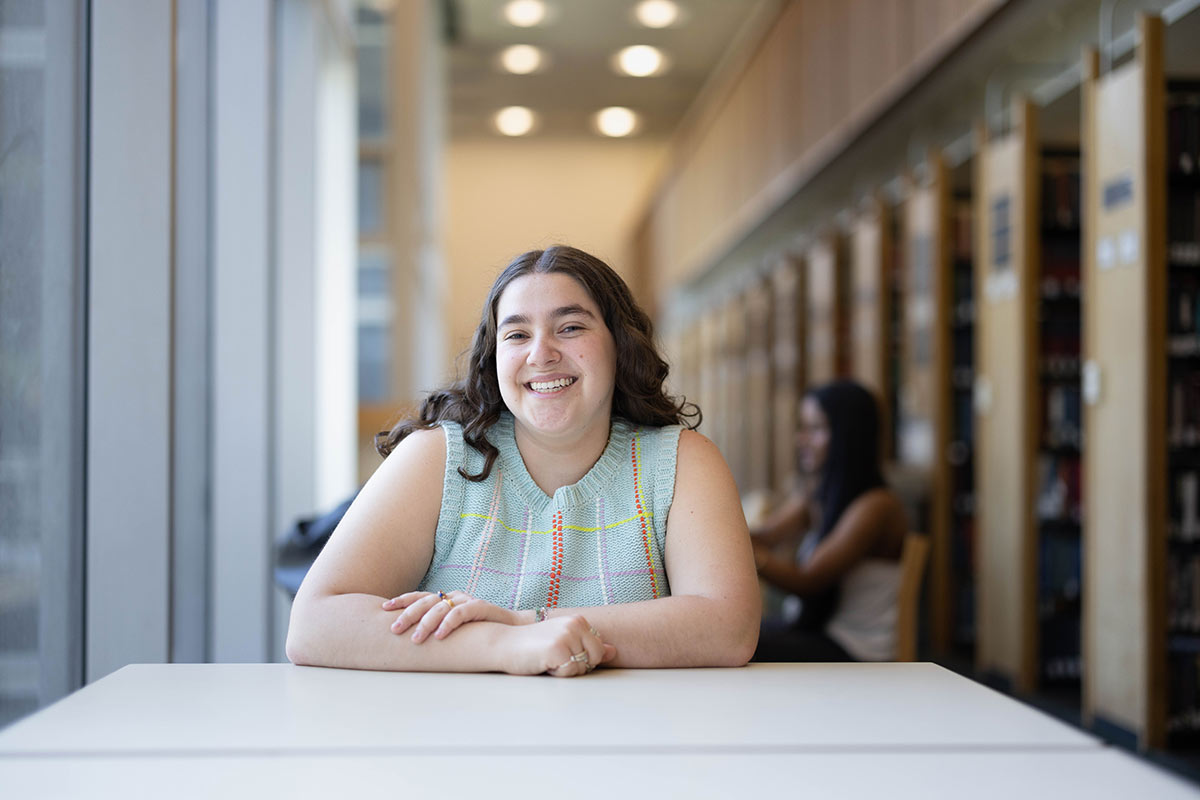 Maia wears a light blue top and sits at a table in the library smiling.