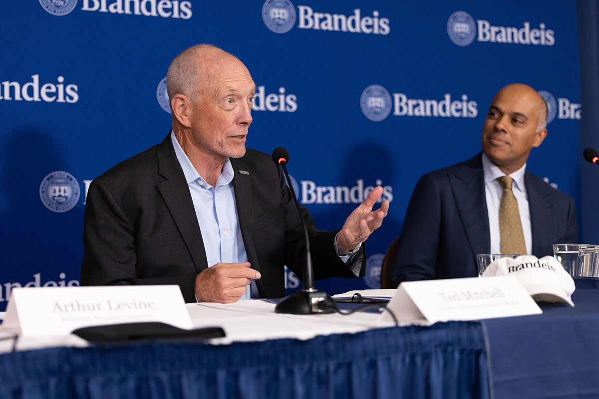 Two panelists sitting and speaking with a Brandeis background behind them