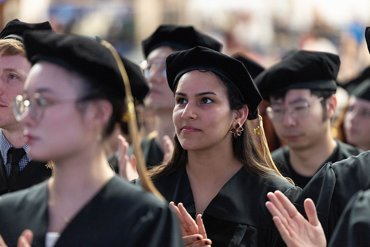 A graduating student sits in the audience at commencement and applauds.