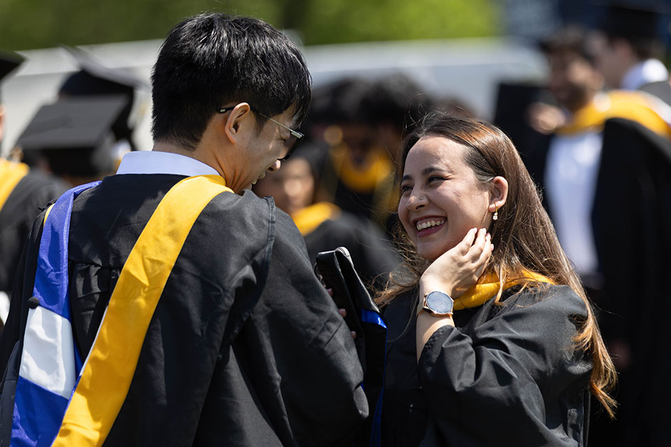 Two students dressed in academic regalia smile and talk outside.