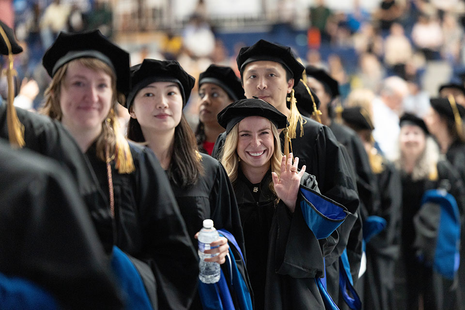 A graduating student smiles and waves at the graduate commencement ceremony.