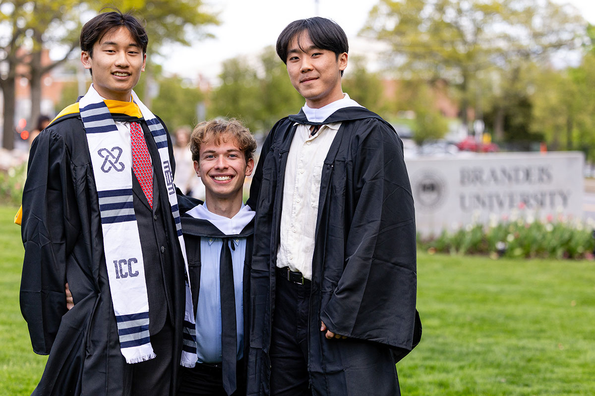 Three students stand together outside on campus dressed in academic regalia.