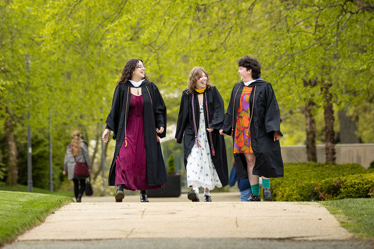 Three students walk through campus dressed in Commencement regalia.
