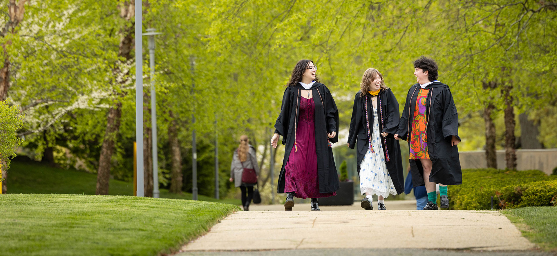 Three students walk through campus dressed in Commencement regalia.