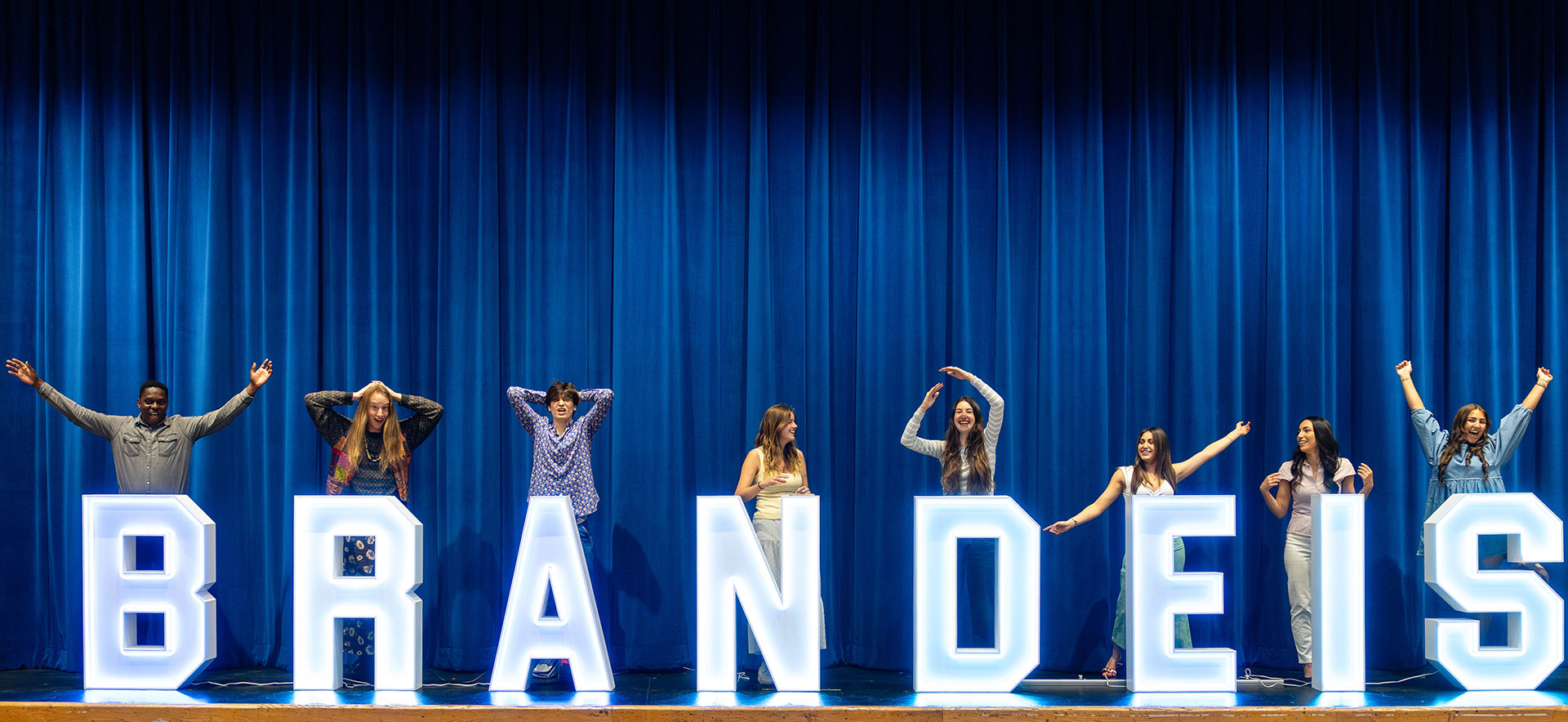 Graduating students stand on a stage behind large letters spelling out the word "Brandeis."