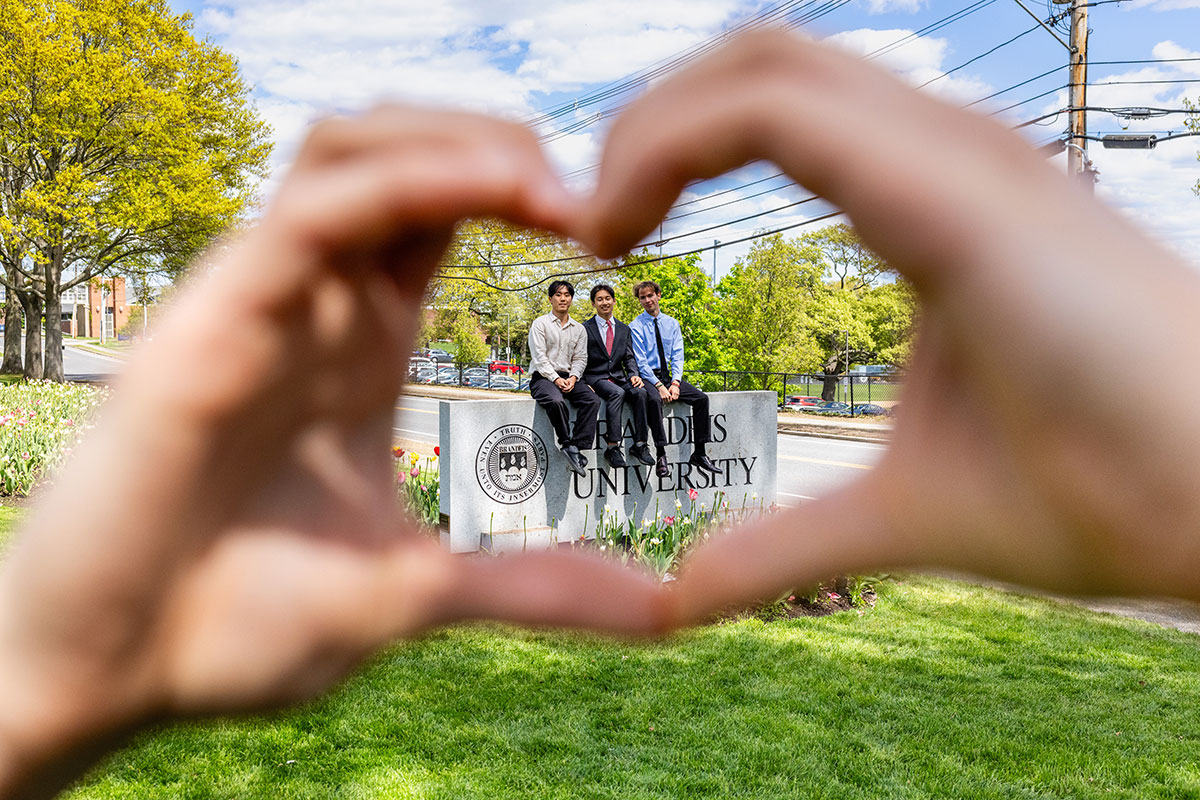 Graduates pose by the Brandeis sign as another student forms a heart with their hands.
