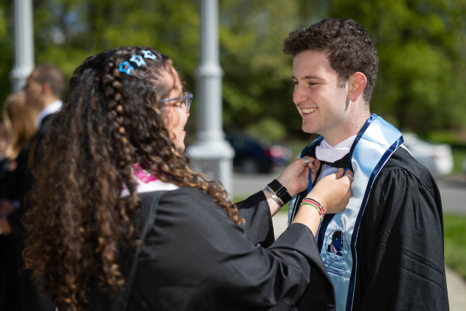 A student adjusts another student's academic regalia.