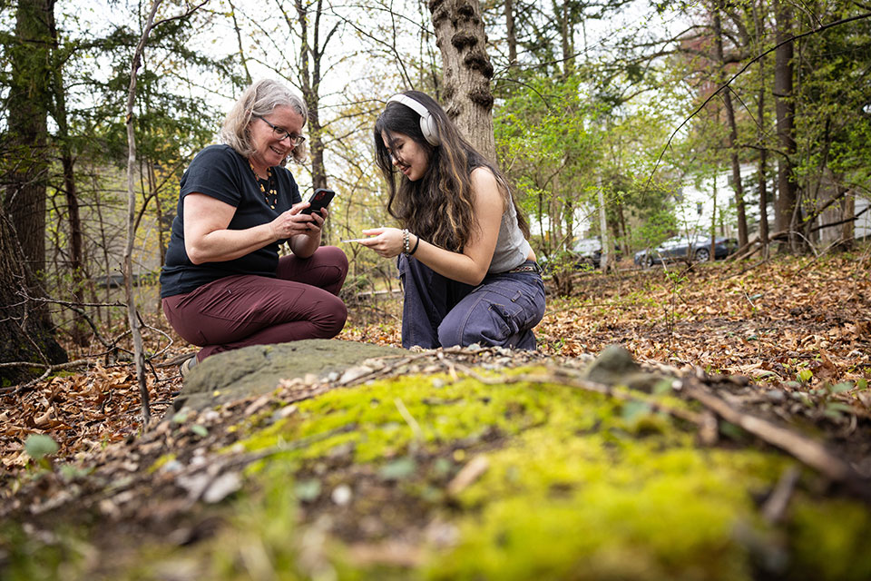 Two people kneel on the ground in the woods taking photos on their phones.