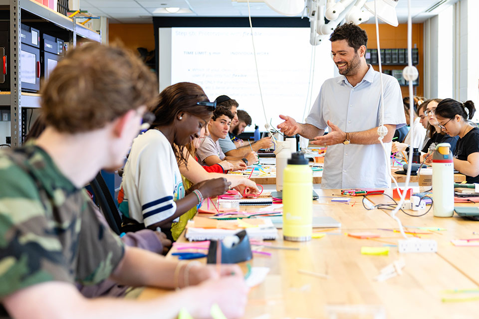 A professor speaks with a classroom of students who are working on a project at a large table.