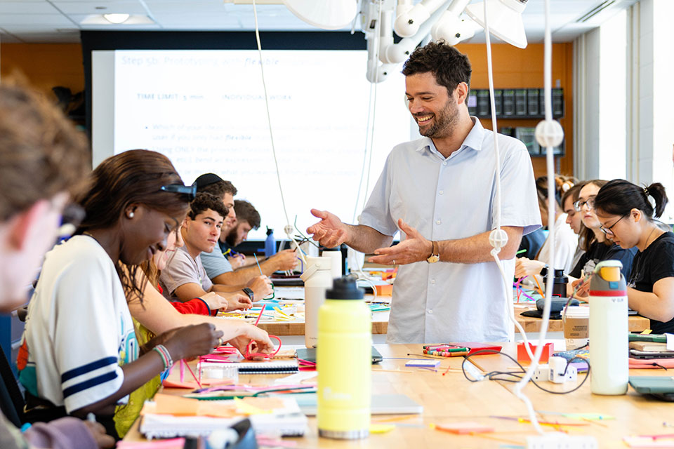 A professor speaks to a classroom of students who are working on a project at a large table.