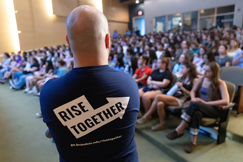 A person looks on to an audience of students wearing a shirt that reads "Rise Together."