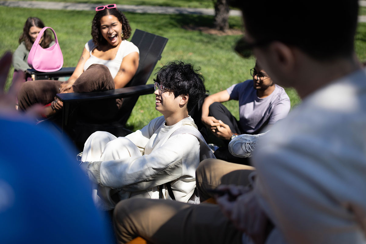 A group of students sit outside on campus and laugh together.