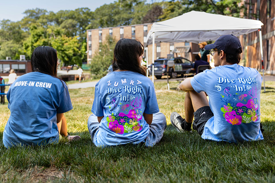 Three students in the move-in crew sit on the grass on campus.