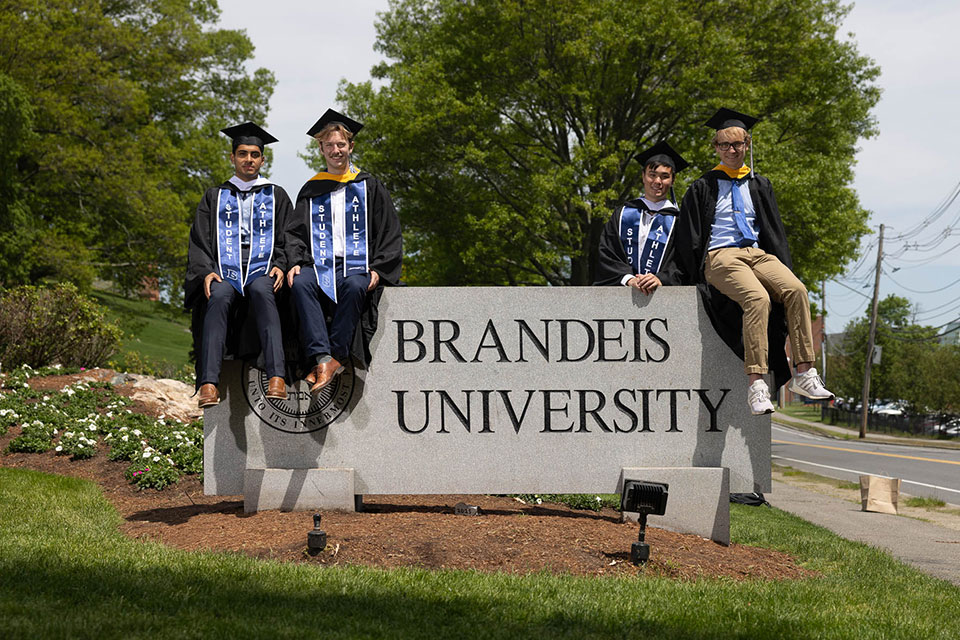 Four students dressed in academic regalia sit on top of the Brandeis University sign before Commencement.