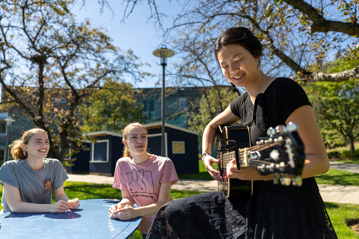 A student plays the guitar for two other students outside on the Brandeis campus.