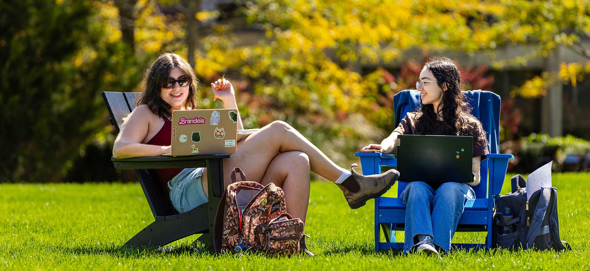Two students sit in chairs on campus working on their laptops.