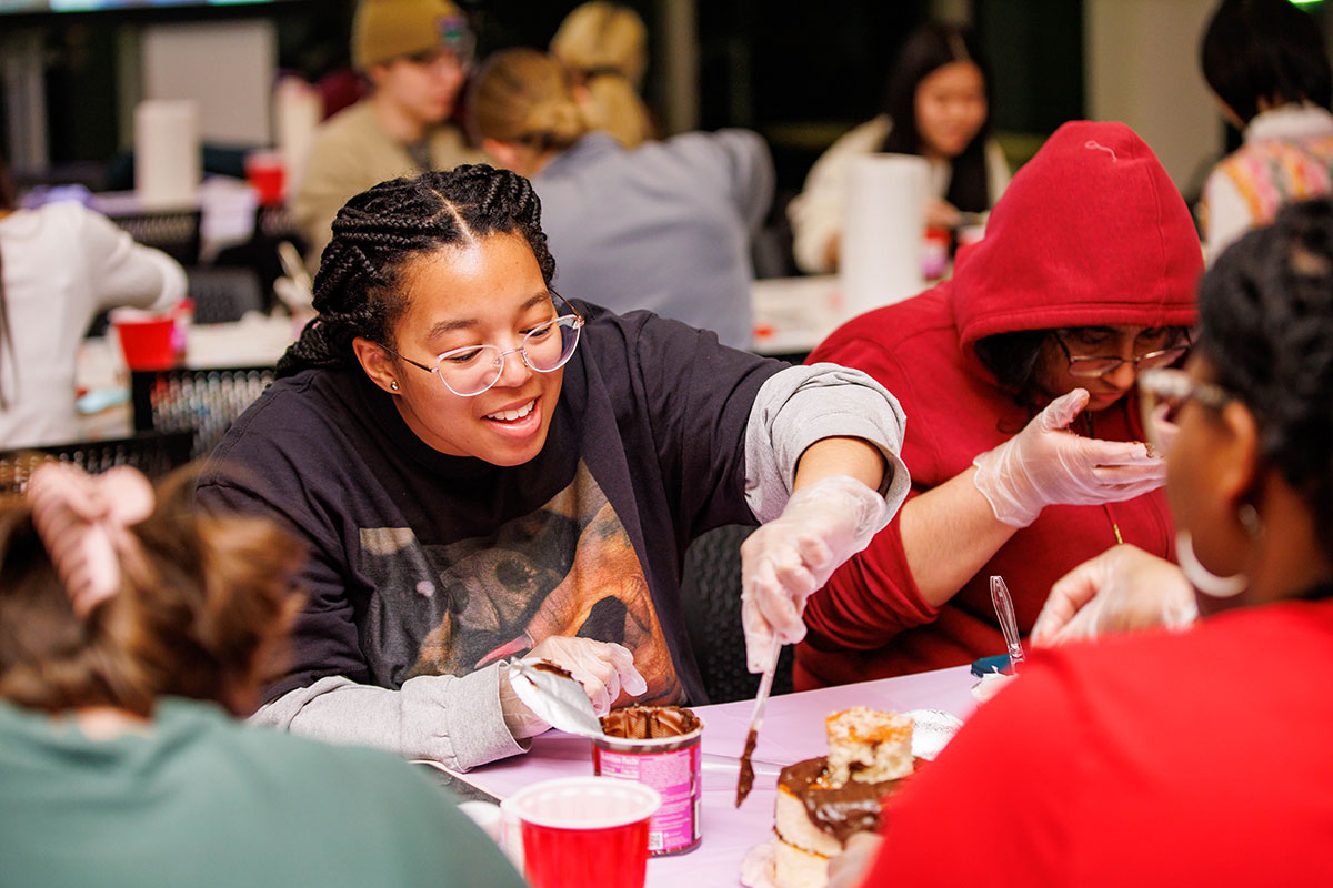 Students sit at a table and decorate cakes with frosting.