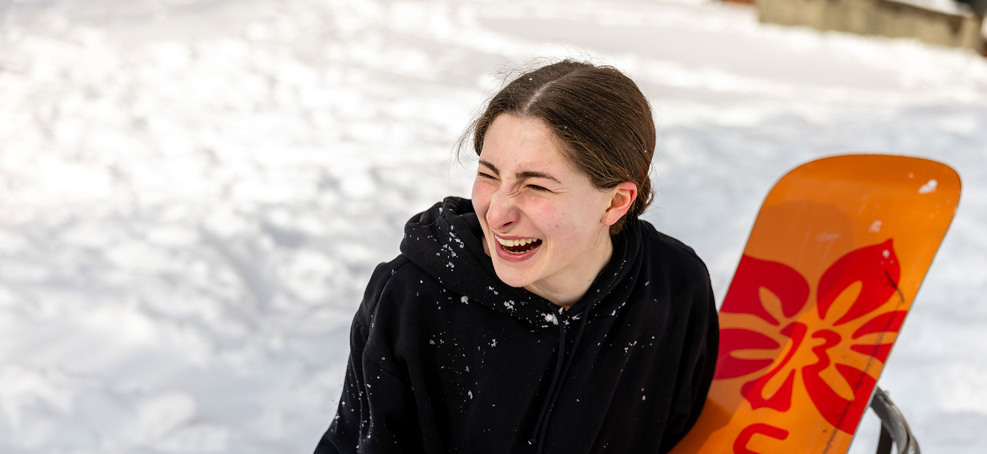 A student holding a snowboard laughs in the snow.