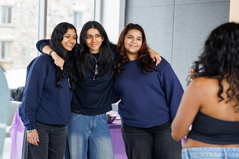 Three students smile with their arms around one another as another student takes their photo.