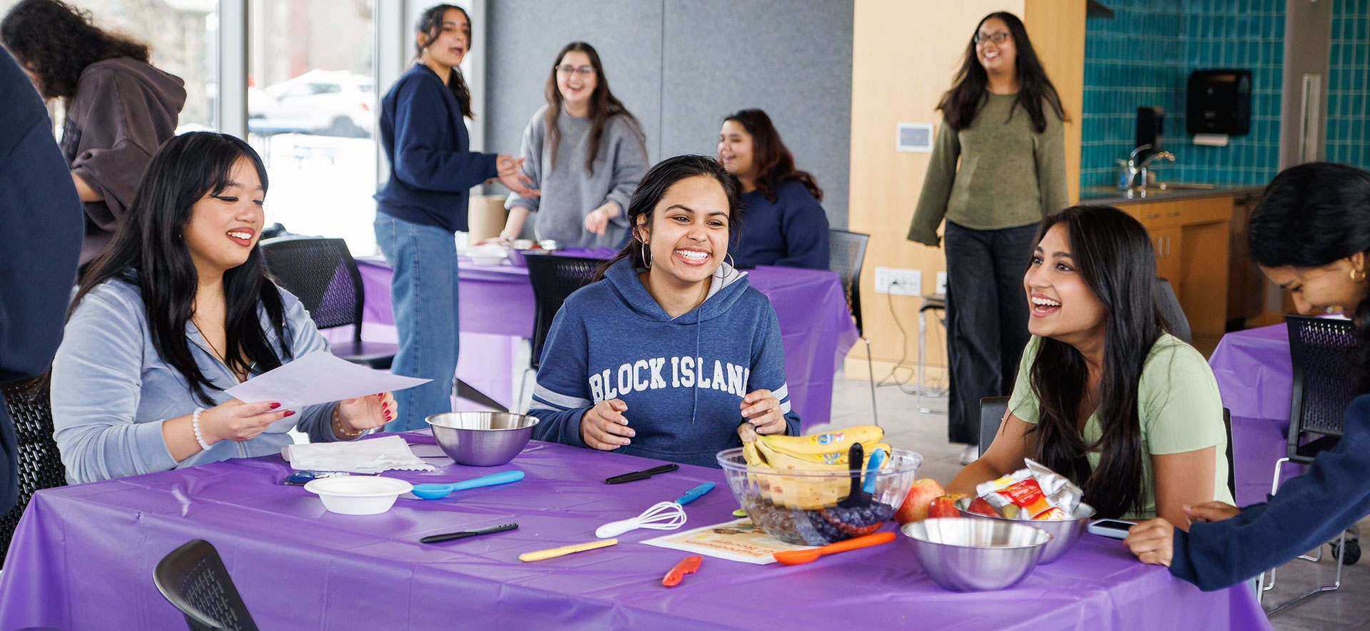 A group of students laugh and sit at a table with ingredients and cooking supplies.